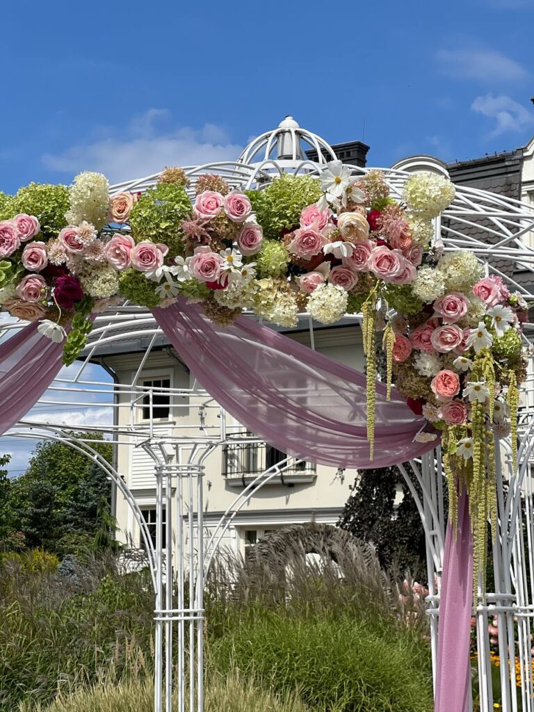 Ślub w plenerze – altana ślubna z dekoracją kwiatową i pudrowymi drapowaniami